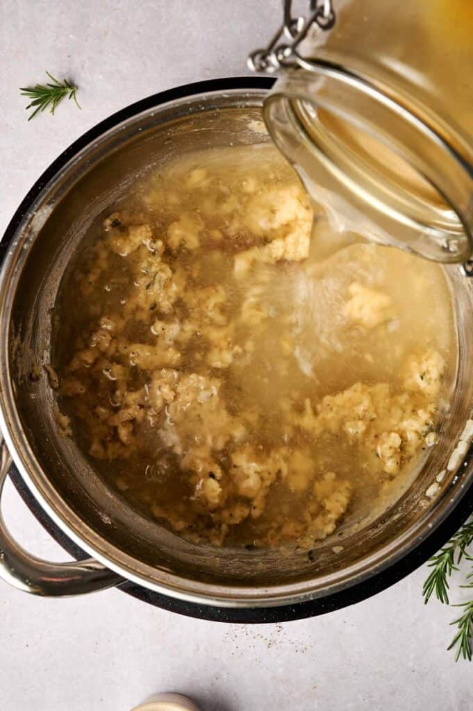 A pot of simmering soup with an open jar pouring turkey gravy into it, surrounded by sprigs of rosemary.