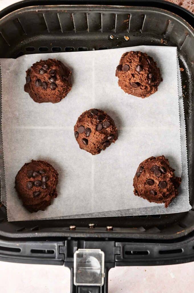 Five decadent copycat Air Fryer Crumbl Chocolate Cake Cookies, studded with rich chocolate chips, rest enticingly on parchment paper inside a sleek black air fryer basket.