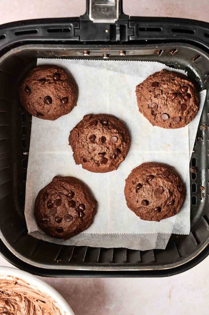 Five copycat Crumbl chocolate cake cookies with chocolate chips rest on parchment paper inside an air fryer basket.