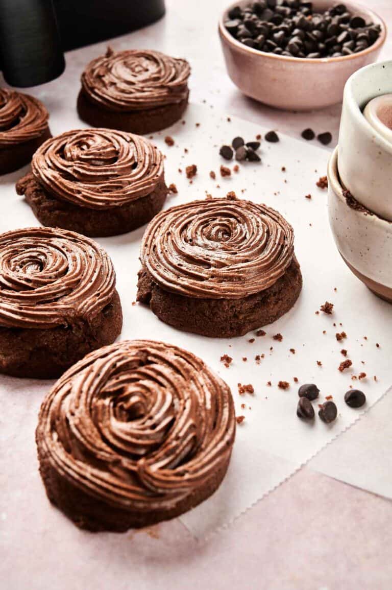 Copycat Crumbl chocolate cake cookies with swirled frosting are arranged on parchment paper. Nearby, two stacked cups and a bowl of chocolate chips add to the cozy scene, inviting you to try this air fryer twist on your favorite treat.