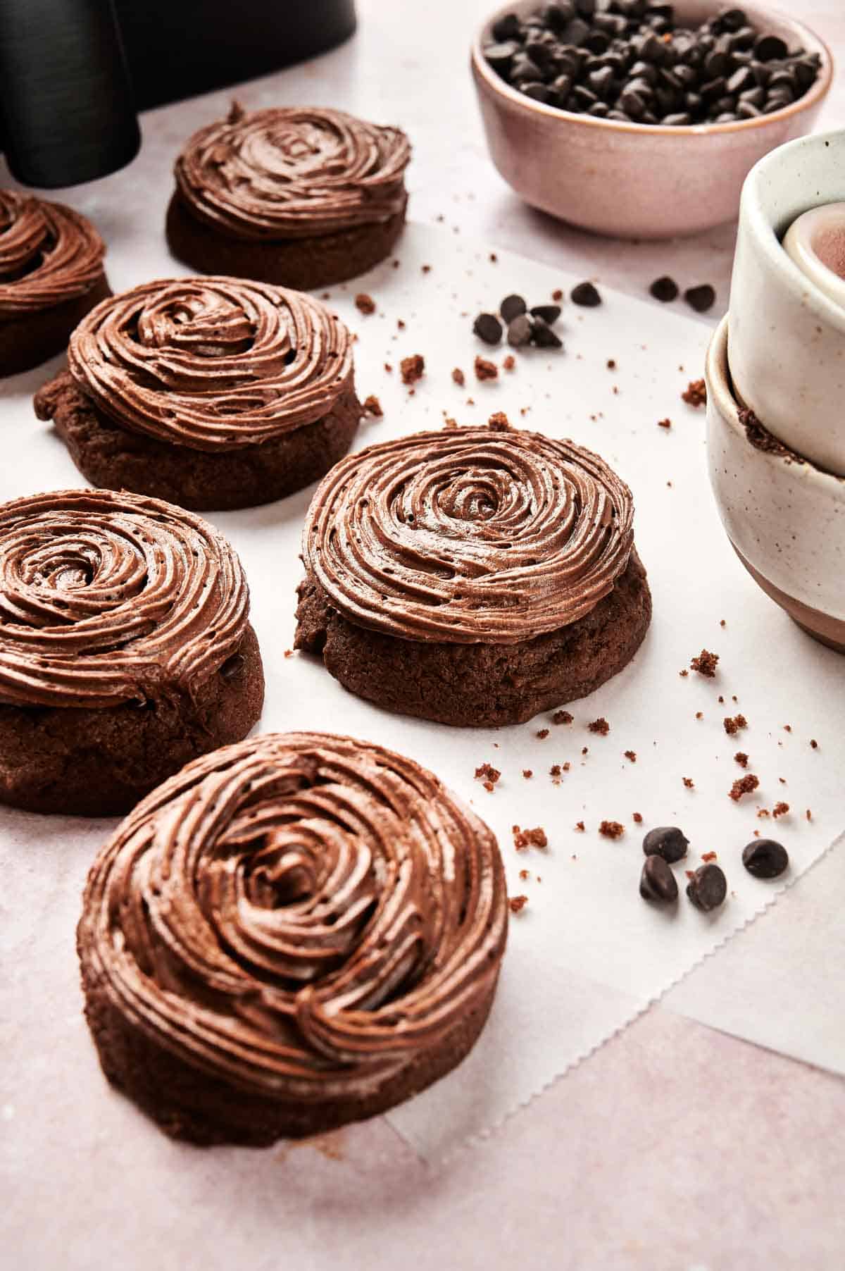 Copycat Crumbl chocolate cake cookies with swirled frosting are arranged on parchment paper. Nearby, two stacked cups and a bowl of chocolate chips add to the cozy scene, inviting you to try this air fryer twist on your favorite treat.