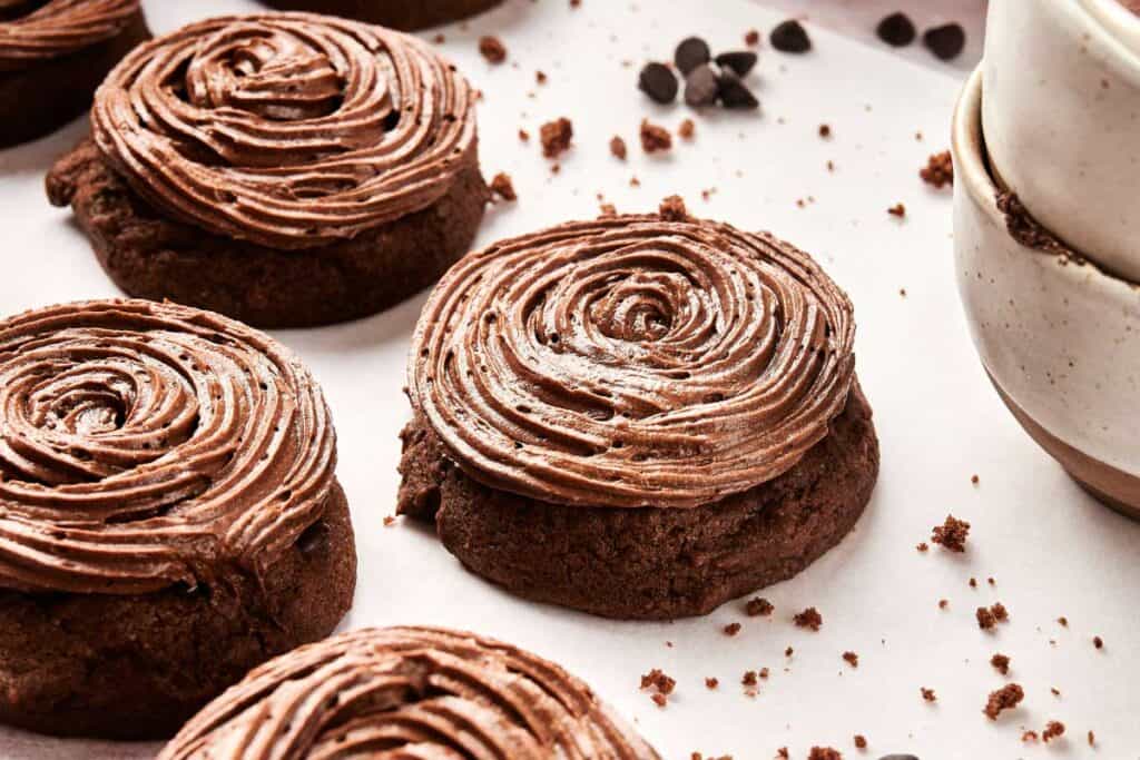 Copycat Crumbl Chocolate Cake Cookies with swirled chocolate frosting rest on parchment paper alongside stacked ceramic bowls and scattered chocolate chips, all conveniently prepared in the air fryer.
