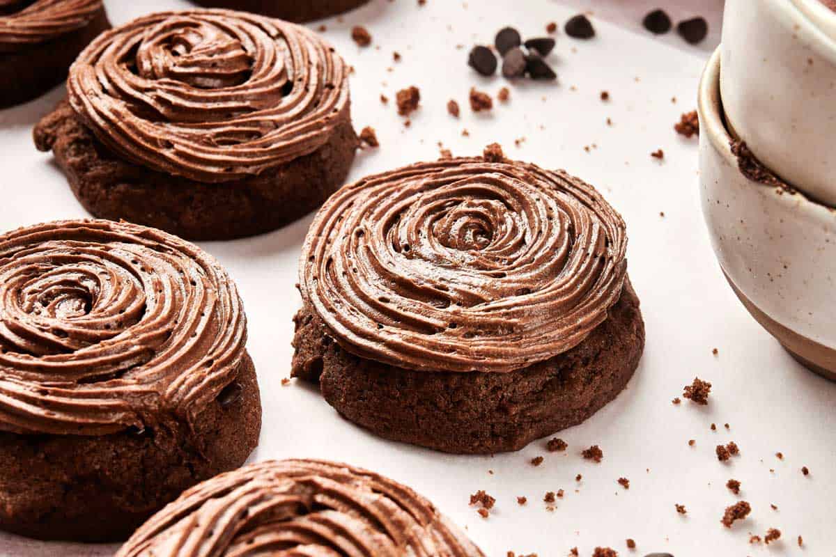 Copycat Crumbl Chocolate Cake Cookies with swirled chocolate frosting rest on parchment paper alongside stacked ceramic bowls and scattered chocolate chips, all conveniently prepared in the air fryer.