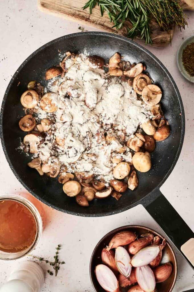 A skillet with mushrooms and flour sits on the countertop, surrounded by shallots, herbs, and a jar of broth&mdash;a perfect start for beef stroganoff.