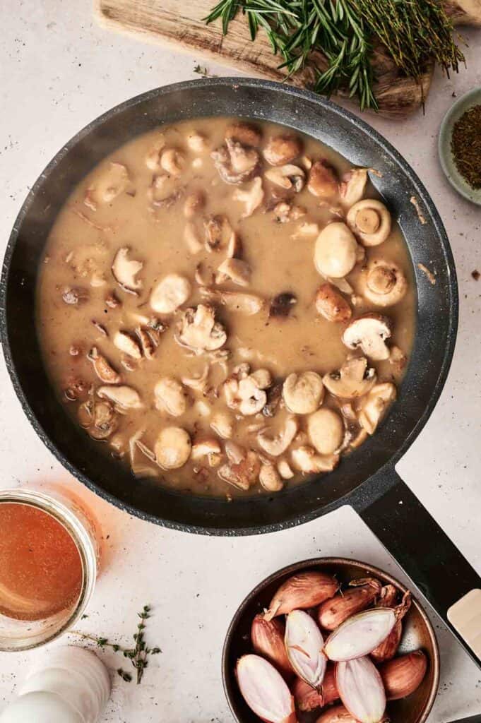 A frying pan filled with creamy mushroom sauce sits on the counter, perfect for topping a classic Beef Stroganoff. Beside it, a glass of liquid, a bowl of halved shallots, and fresh herbs are ready to enhance the dish's rich flavors.