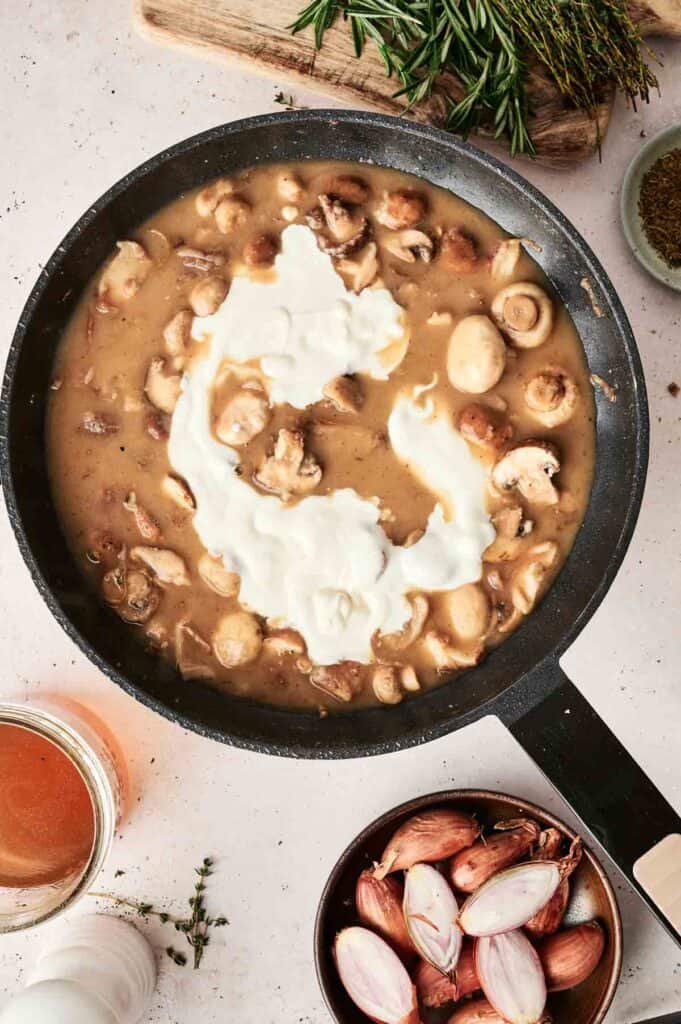 A skillet with Beef Stroganoff mushroom sauce and cream being mixed, surrounded by shallots, herbs, and a beverage on a kitchen counter.