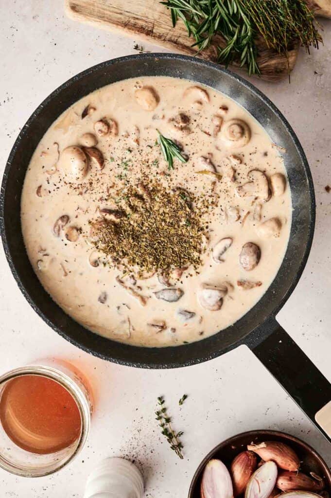 Pan of creamy mushroom soup garnished with herbs, reminiscent of a delicate Beef Stroganoff, surrounded by cooking ingredients like shallots, savory herbs, and rich broth on a light background.