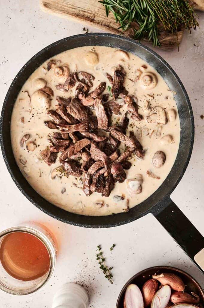 A pan of creamy mushroom sauce with sliced beef, reminiscent of classic Beef Stroganoff, surrounded by shallots, herbs, and a glass of broth on a light surface.