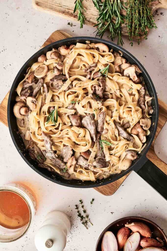 A skillet of creamy Beef Stroganoff pasta with mushrooms and herbs rests on a wooden board. Nearby, a drink accompanies scattered herbs and shallots on the white surface.