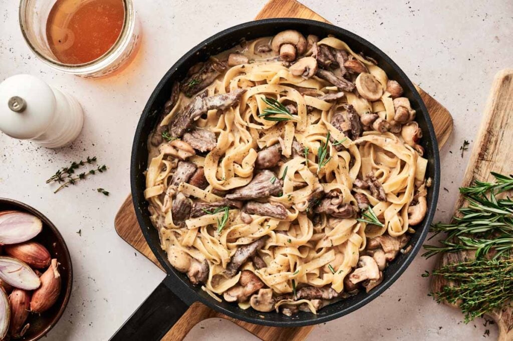 A pan of creamy Beef Stroganoff pasta with mushrooms and tender meat, garnished with fresh rosemary, sits on a wooden board next to shallots, herbs, a drink, and a pepper grinder.