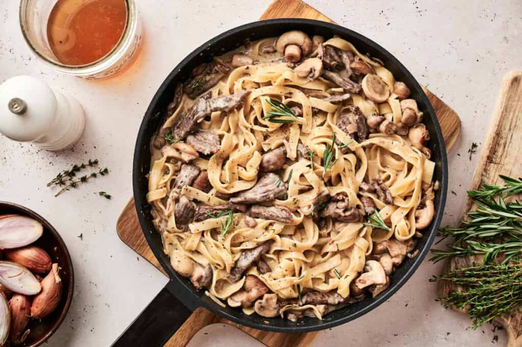Pan of creamy pasta with mushrooms and herbs on a wooden board, reminiscent of Beef Stroganoff, accompanied by a glass of drink, rosemary, shallots, and a pepper mill on a light textured surface.