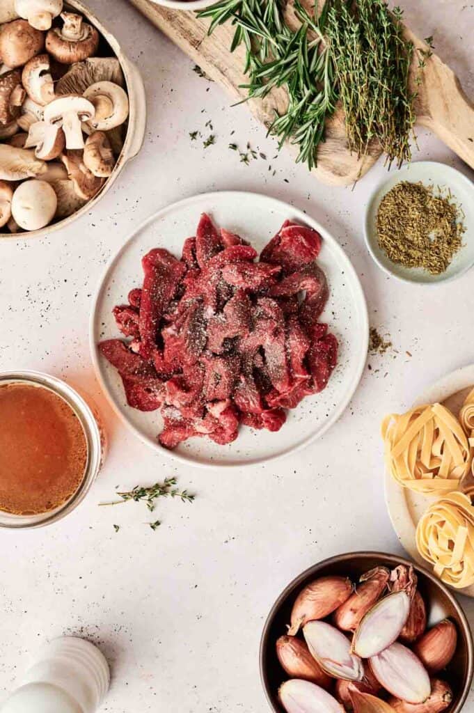 A plate of raw beef strips seasoned with salt and pepper, reminiscent of a deconstructed Beef Stroganoff, is surrounded by mushrooms, herbs, pasta, broth, and shallots on a light-colored surface.