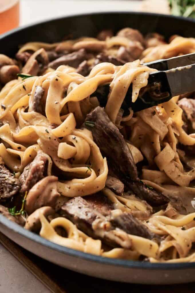 A pan filled with creamy fettuccine pasta and slices of beef stroganoff is being expertly stirred with tongs.