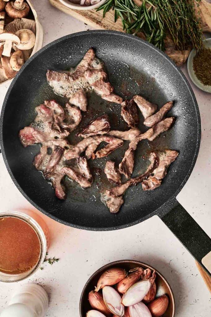 Slices of beef sizzle in a frying pan, resembling the start of a delicious Beef Stroganoff, surrounded by mushrooms, shallots, and herbs on the countertop.