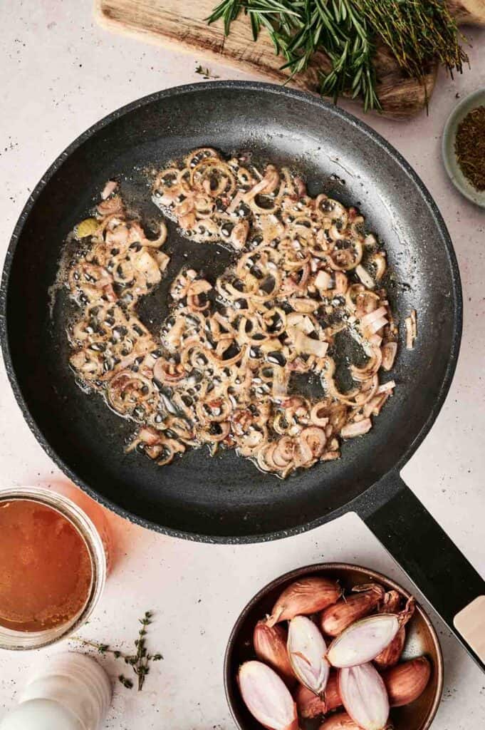 Chopped shallots saut&eacute;ing in a black pan, with a bowl of halved shallots, fresh herbs, and a cup of broth nearby, set the stage for crafting rich and creamy Beef Stroganoff.
