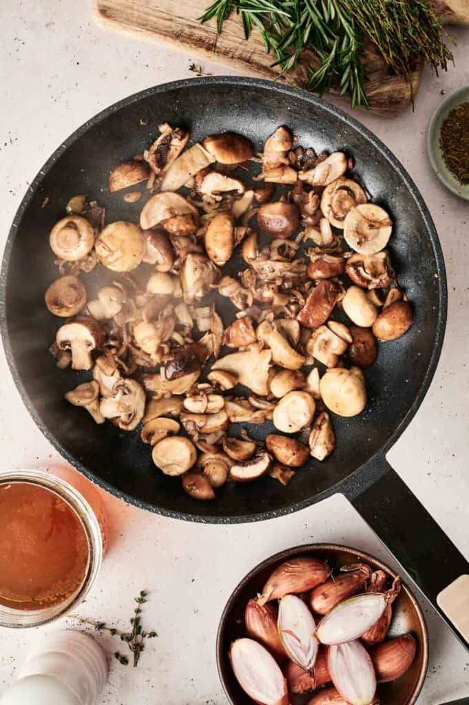 A pan with saut&eacute;ed mushrooms, reminiscent of a cozy Beef Stroganoff recipe, rests on a light surface, surrounded by ingredients like a bowl of shallots, herbs, and a jar of liquid.