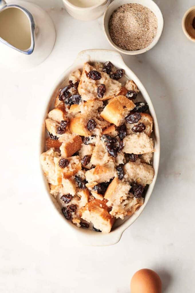 A baking dish filled with delectable bread pudding topped with raisins takes center stage, surrounded by bowls of milk, cinnamon sugar, and an egg on a marble surface.