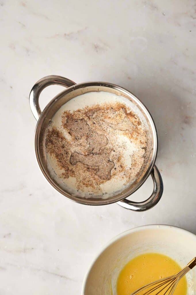 A saucepan with milk and spices, likely cinnamon, suggests the start of a delightful bread pudding. Below, a bowl contains a whisk and a yellow mixture, possibly eggs, sitting on the marble surface.
