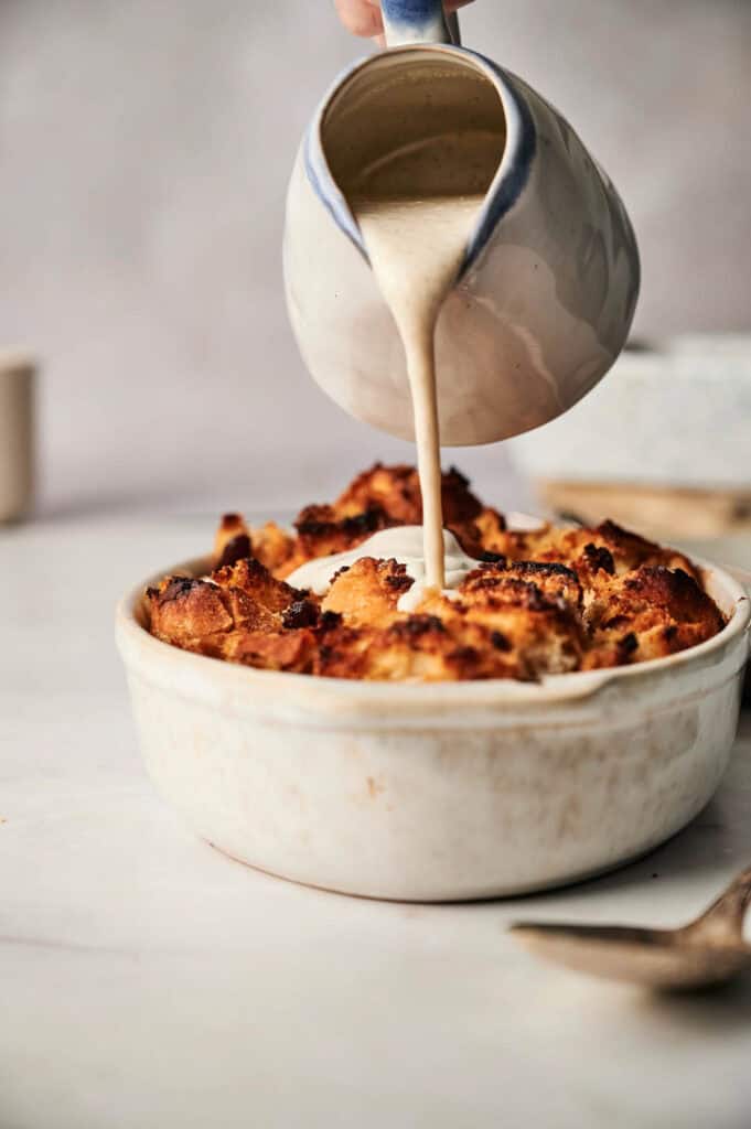 A luscious creamy sauce cascades from a jug, enveloping the warm bread pudding nestled in its round ceramic dish.