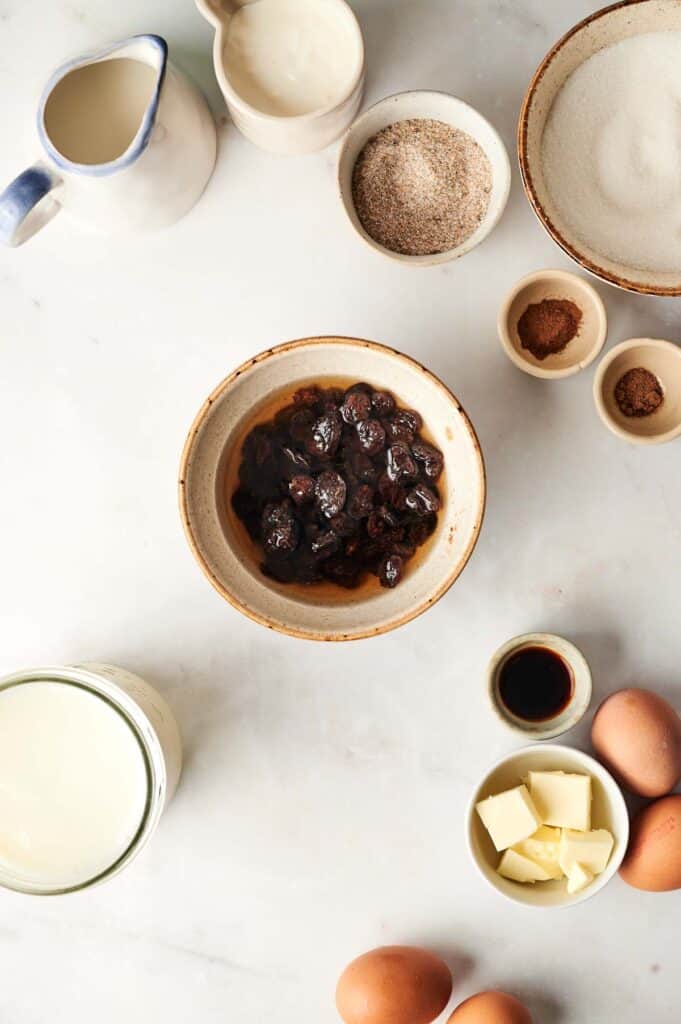 Ingredients for bread pudding laid out on a countertop include milk, butter, eggs, sugar, spices, flour, a bowl of dark fruit, and a jug of cream.