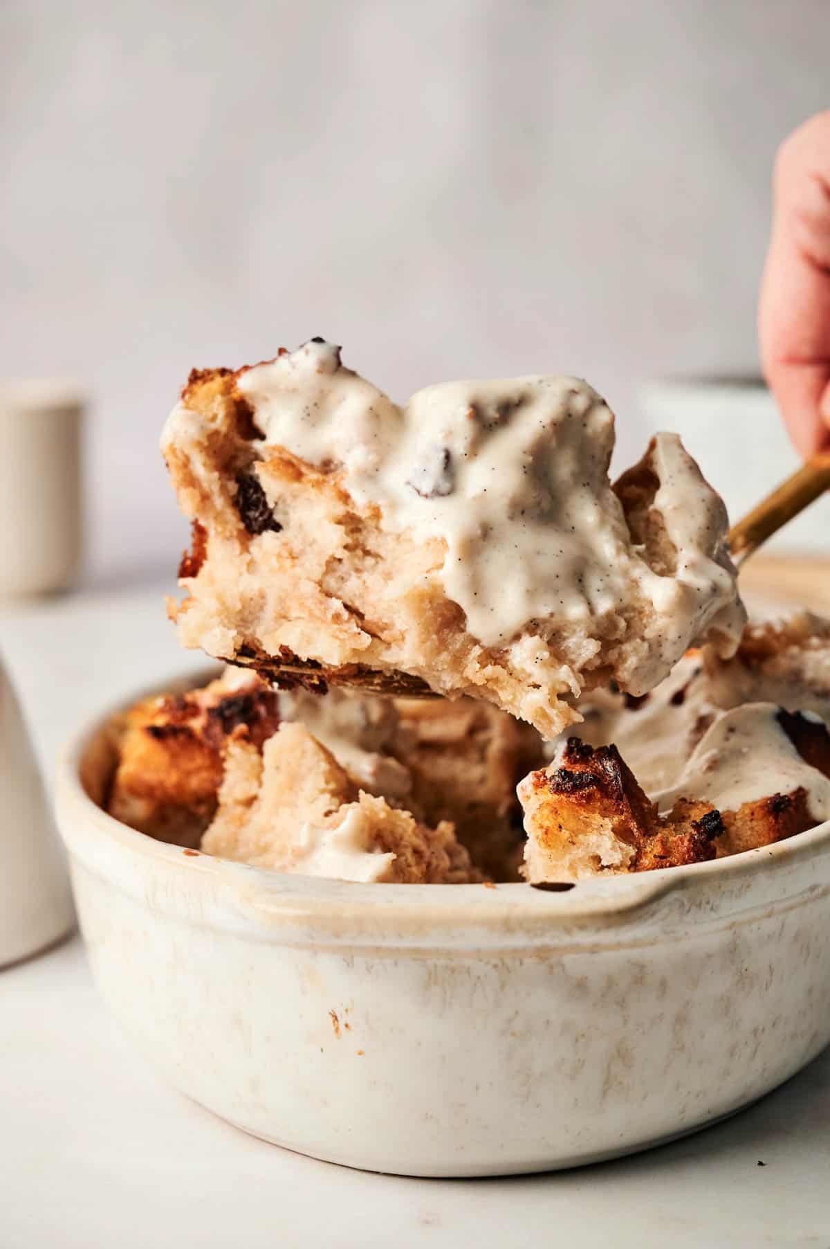 A spoon gracefully scoops a luscious serving of warm bread pudding topped with rich, creamy vanilla sauce from a pristine white bowl.