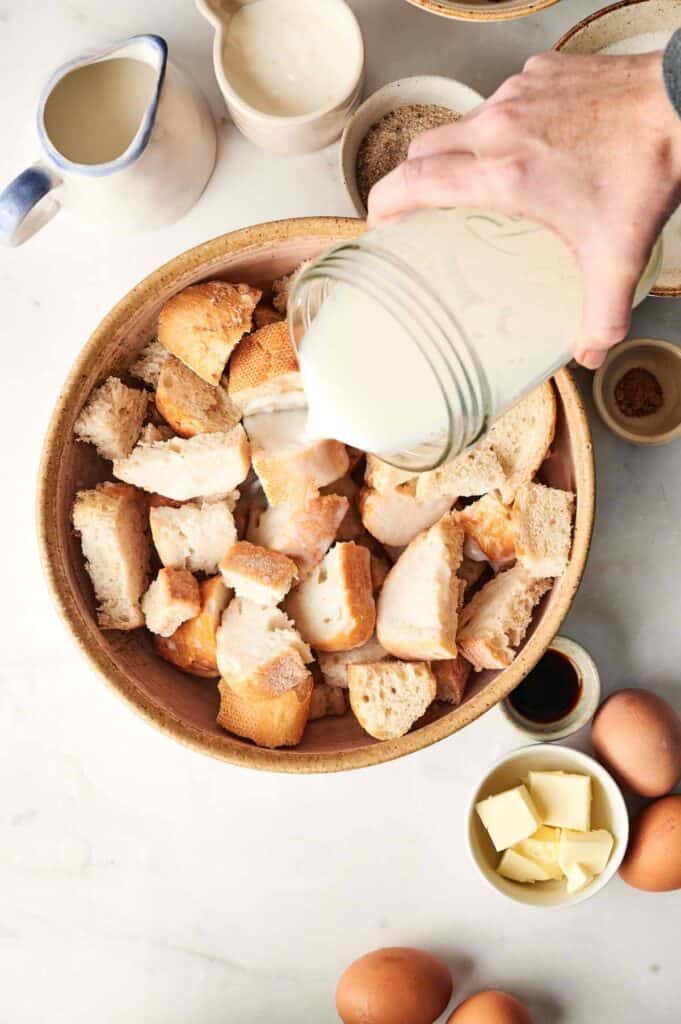 A hand pours milk over a bowl filled with torn bread pieces, crafting the beginnings of a luscious bread pudding. Surrounding the bowl are eggs, butter cubes, spices, and a jug.