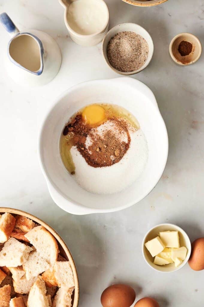 The ingredients for a delectable bread pudding rest on the marble countertop: a white mixing bowl filled with eggs, sugar, and spices, surrounded by a pitcher of milk, cream, cubed bread, and butter.