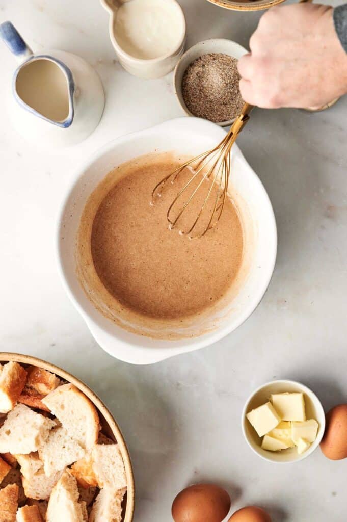 A person is whisking brown batter in a white bowl on a marble surface, crafting the perfect bread pudding, surrounded by ingredients like bread pieces, butter cubes, eggs, and a pitcher of cream.