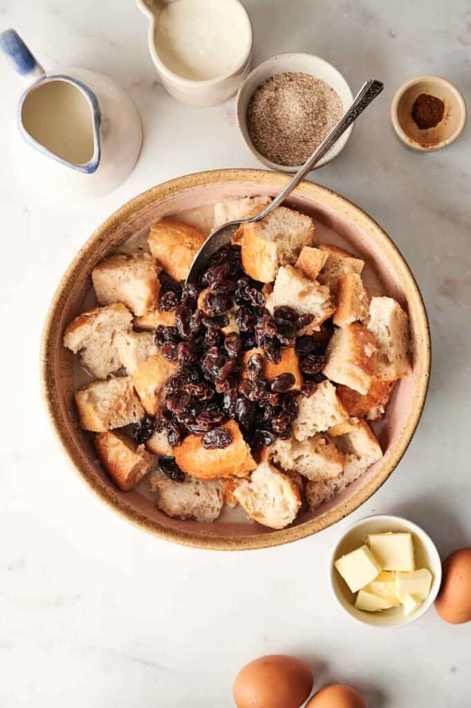 A bowl of bread pudding cubes topped with raisins, accompanied by a pitcher, cream, sugar, cinnamon, eggs, and butter. A spoon rests in the bowl.
