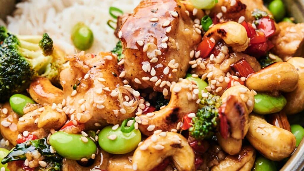Close-up of a dish with chicken, broccoli, cashews, edamame, red peppers, and sesame seeds served over rice.