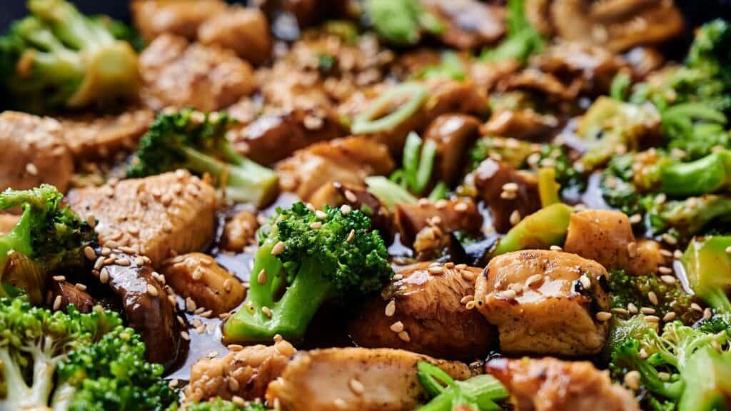Close-up of a stir-fry dish featuring chicken, broccoli, and mushrooms, garnished with sesame seeds.