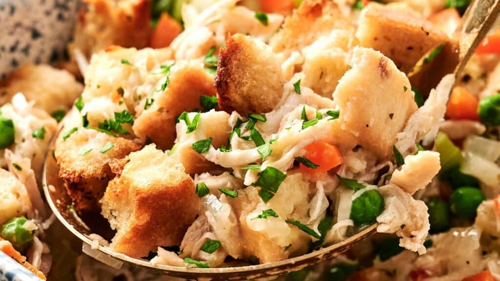 A close-up of a spoonful of chicken pot pie casserole with chunks of bread, peas, carrots, shredded chicken, and chopped parsley.