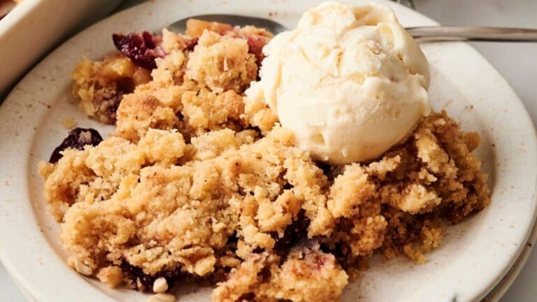 A plate of fruit crumble topped with a scoop of vanilla ice cream on a white plate. A spoon rests beside the dessert.