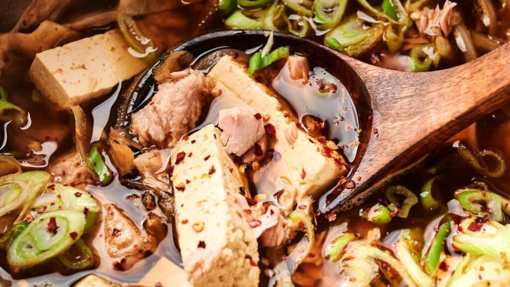 A close-up of a wooden spoon in a bowl of soup with tofu, green onions, meat, and chili flakes.