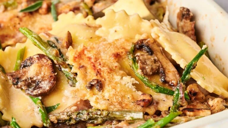 Close-up of baked ravioli with mushrooms, asparagus, and grated cheese in a creamy sauce.