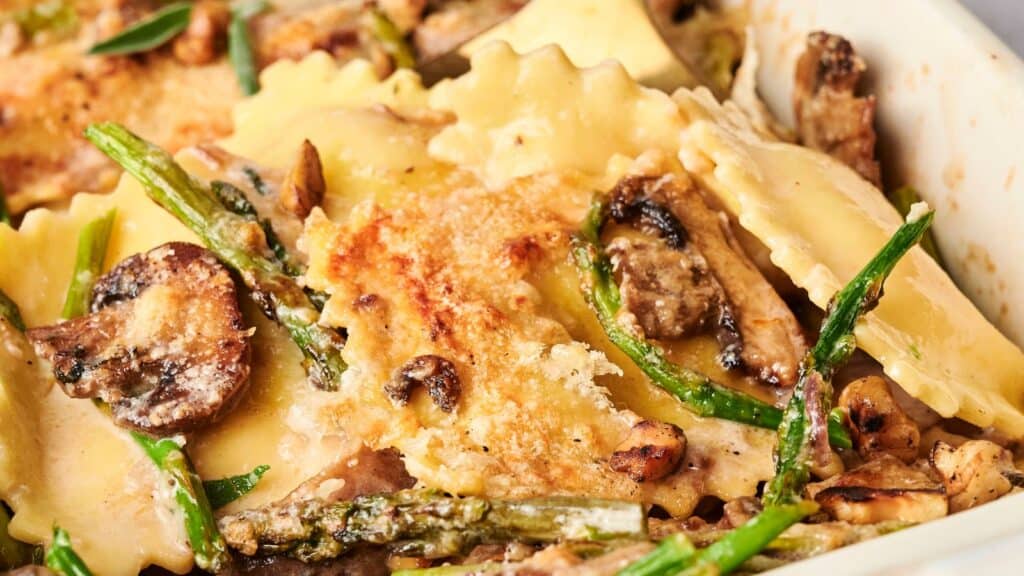 Close-up of a baked dish with ravioli, mushrooms, asparagus, and melted cheese on top.