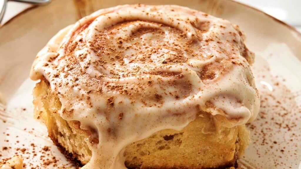 Close-up of a cinnamon roll with cream cheese frosting, sprinkled with cinnamon on a beige plate.