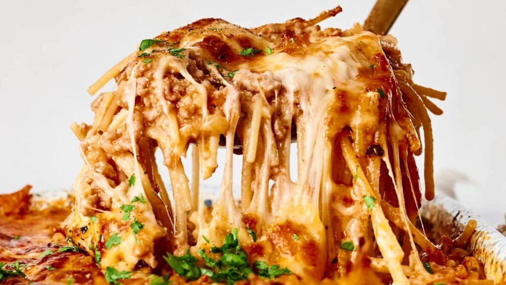 A close-up of cheesy baked spaghetti being lifted from a casserole dish, showing melted cheese and garnish on top.