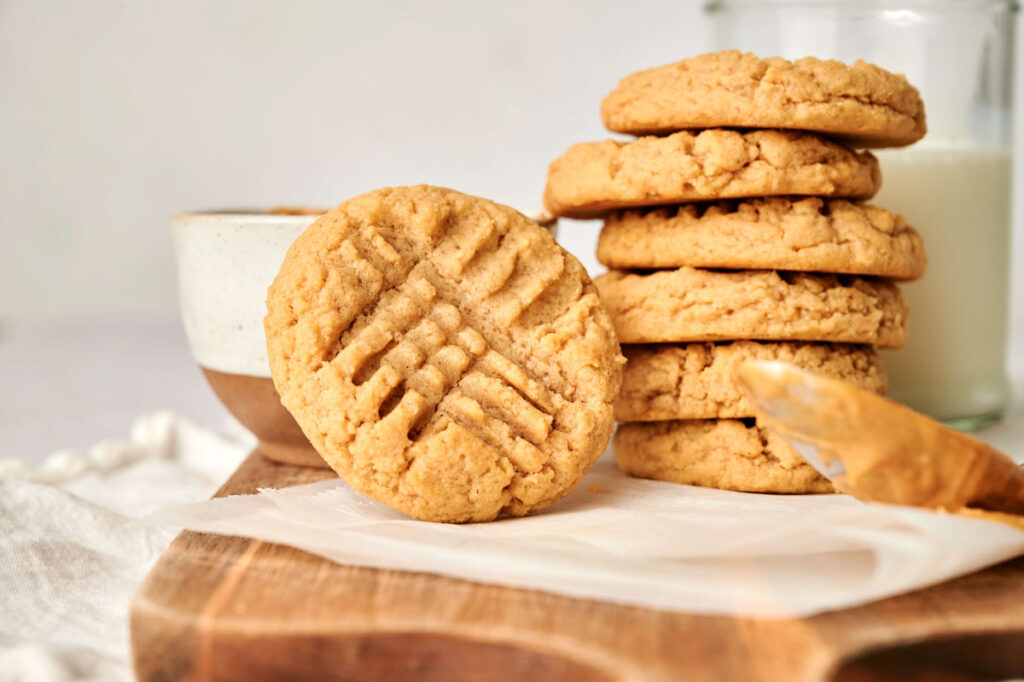 A stack of delicious Peanut Butter Cookies sits on a wooden board, with one cookie placed invitingly in front. In the background, a jar of milk and a bowl complete this classic scene.
