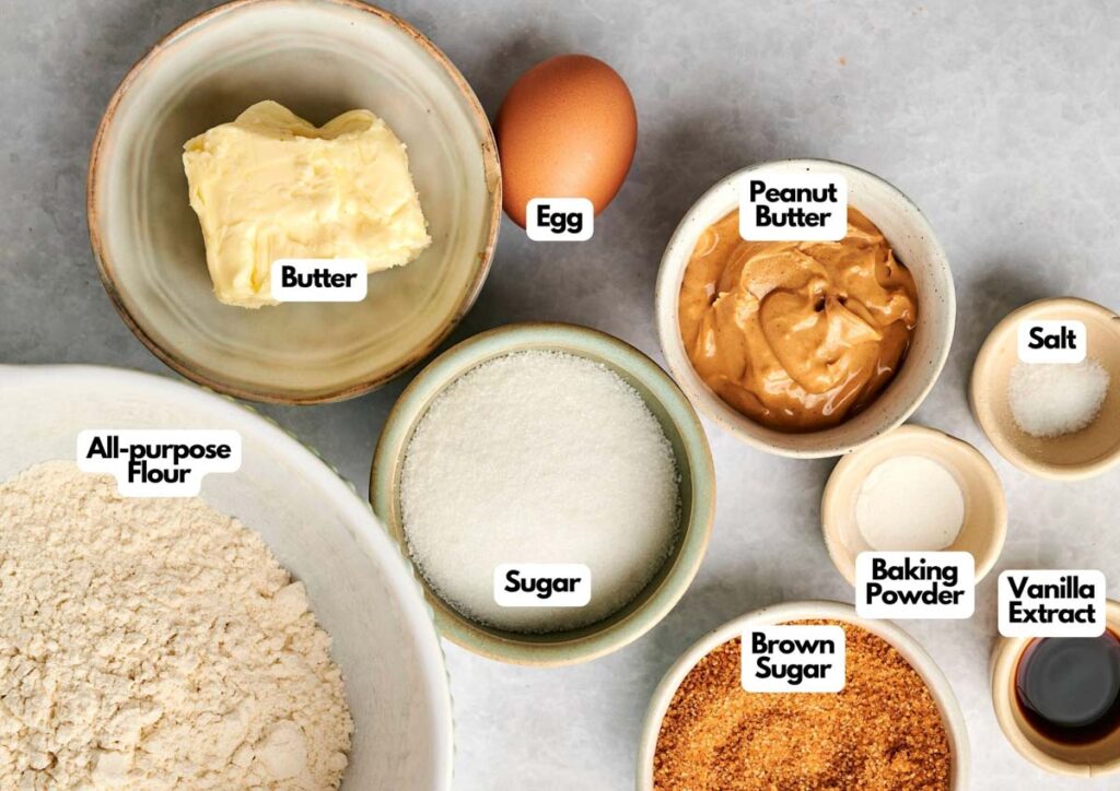 Bowls containing butter, an egg, peanut butter, salt, all-purpose flour, sugar, baking powder, vanilla extract, and brown sugar are arranged on a countertop—ready for making classic peanut butter cookies.