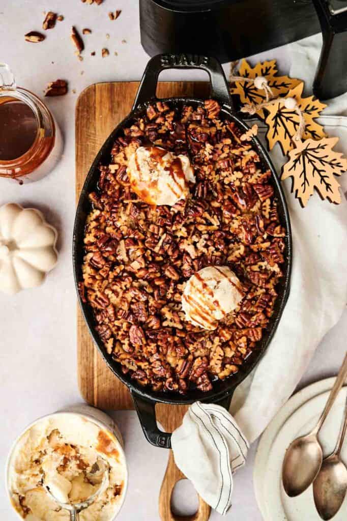 Top view of an Air Fryer Pecan Pie Cobbler dessert topped with ice cream and caramel drizzle in a black baking dish, surrounded by autumn-themed decorations, utensils, and a small pumpkin.