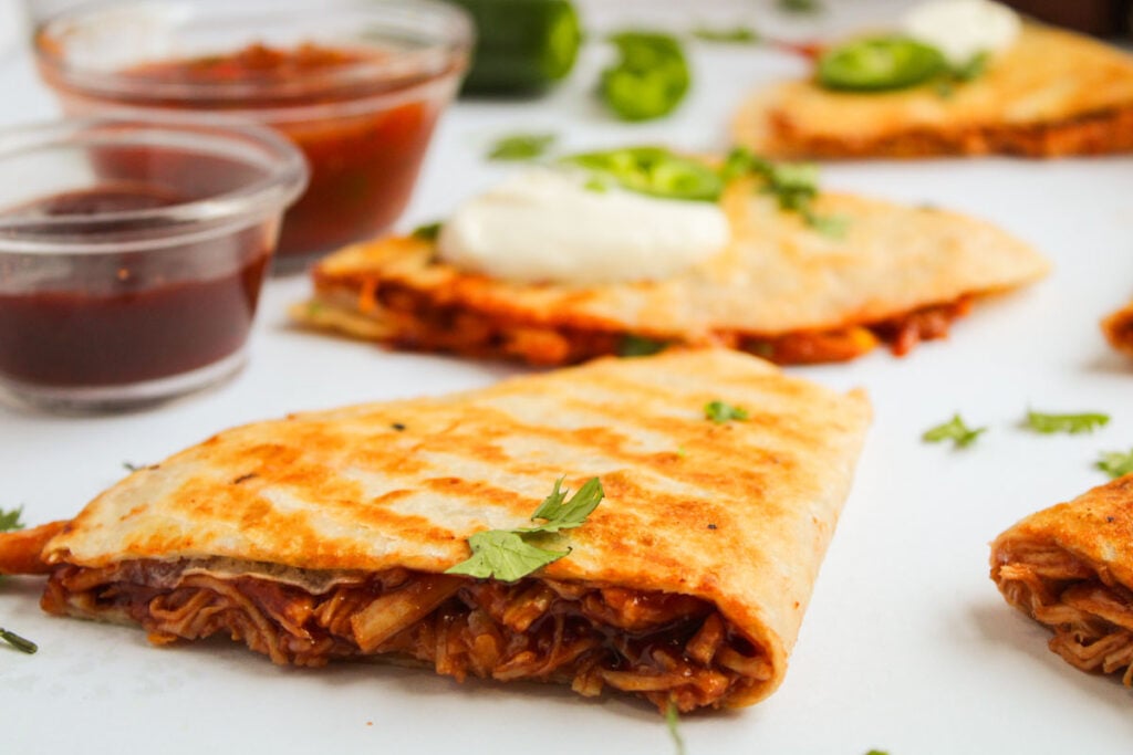 A close-up of a BBQ Chicken Quesadilla wedge topped with sour cream and cilantro, with salsa and barbecue sauce in small cups in the background.