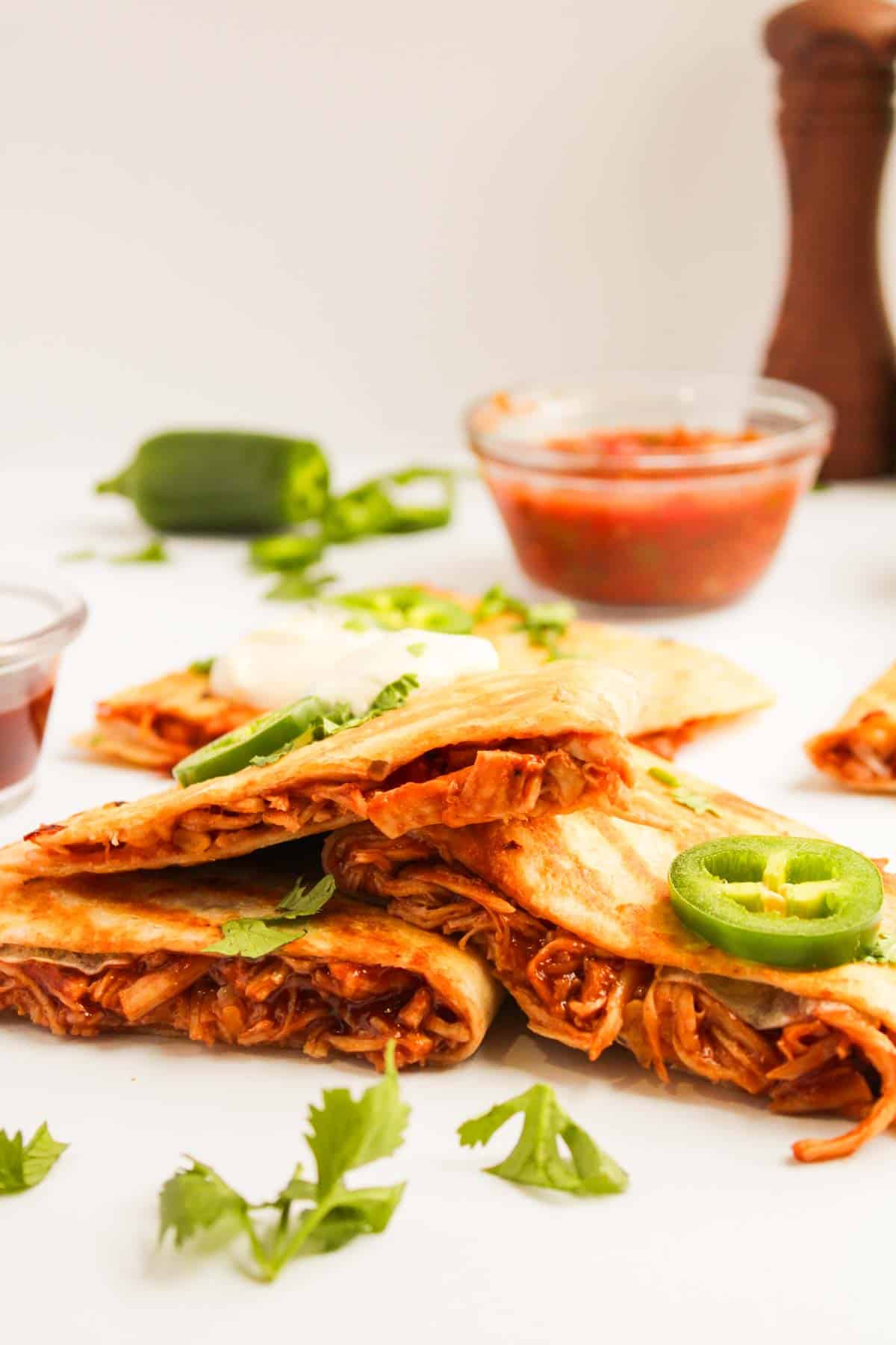 Four BBQ chicken quesadilla wedges filled with shredded meat and topped with jalapeño slices are arranged on a white surface. Salsa and cilantro garnish are in the background.