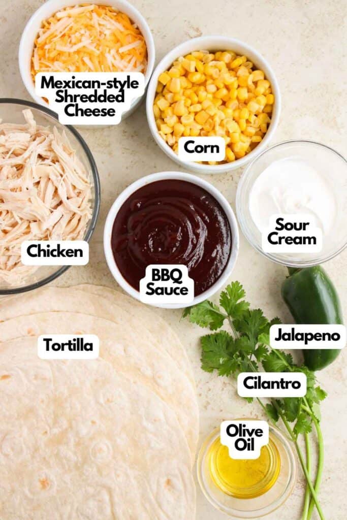 Ingredients for a BBQ Chicken Quesadilla including shredded cheese, corn, chicken, BBQ sauce, sour cream, tortilla, cilantro, jalapeño, and olive oil are beautifully displayed in bowls and on a surface.