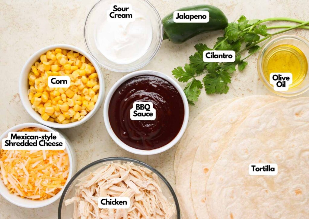 Ingredients laid out for a BBQ Chicken Quesadilla recipe: corn, sour cream, jalapeno, cilantro, olive oil, tortillas, shredded chicken, BBQ sauce, and Mexican-style shredded cheese.