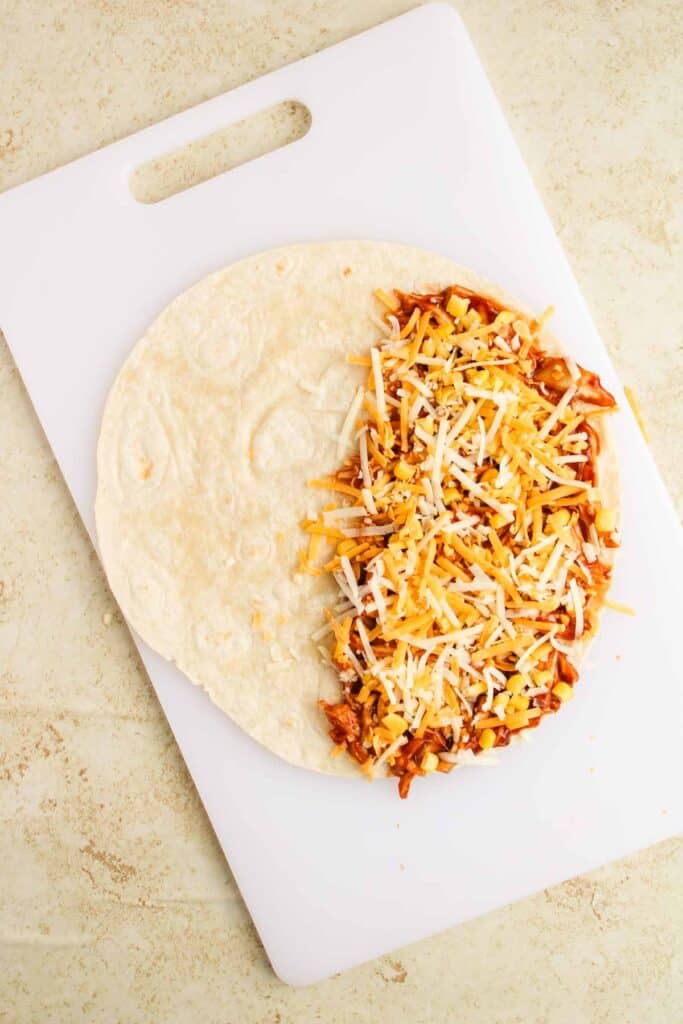 A BBQ Chicken Quesadilla sits invitingly on a white cutting board, its tortilla half-filled with shredded cheese and tender shredded chicken.