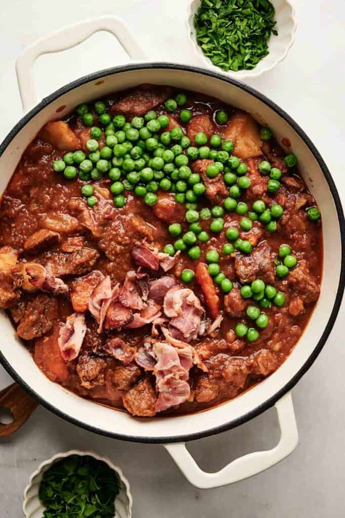 A white pot filled with beef stew, topped with peas and bacon pieces. Small bowls of chopped parsley surround the pot.
