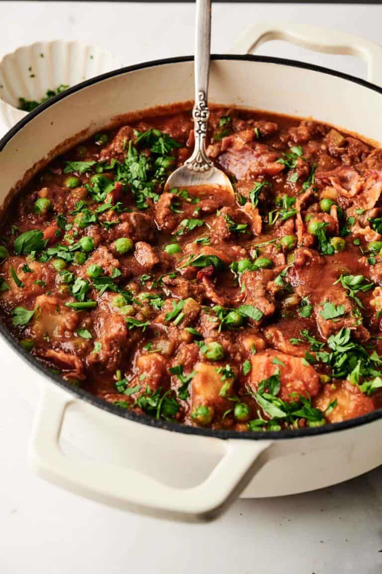 A white pot filled with a hearty beef stew, featuring chunks of meat, peas, carrots, and herbs, with a spoon resting inside.