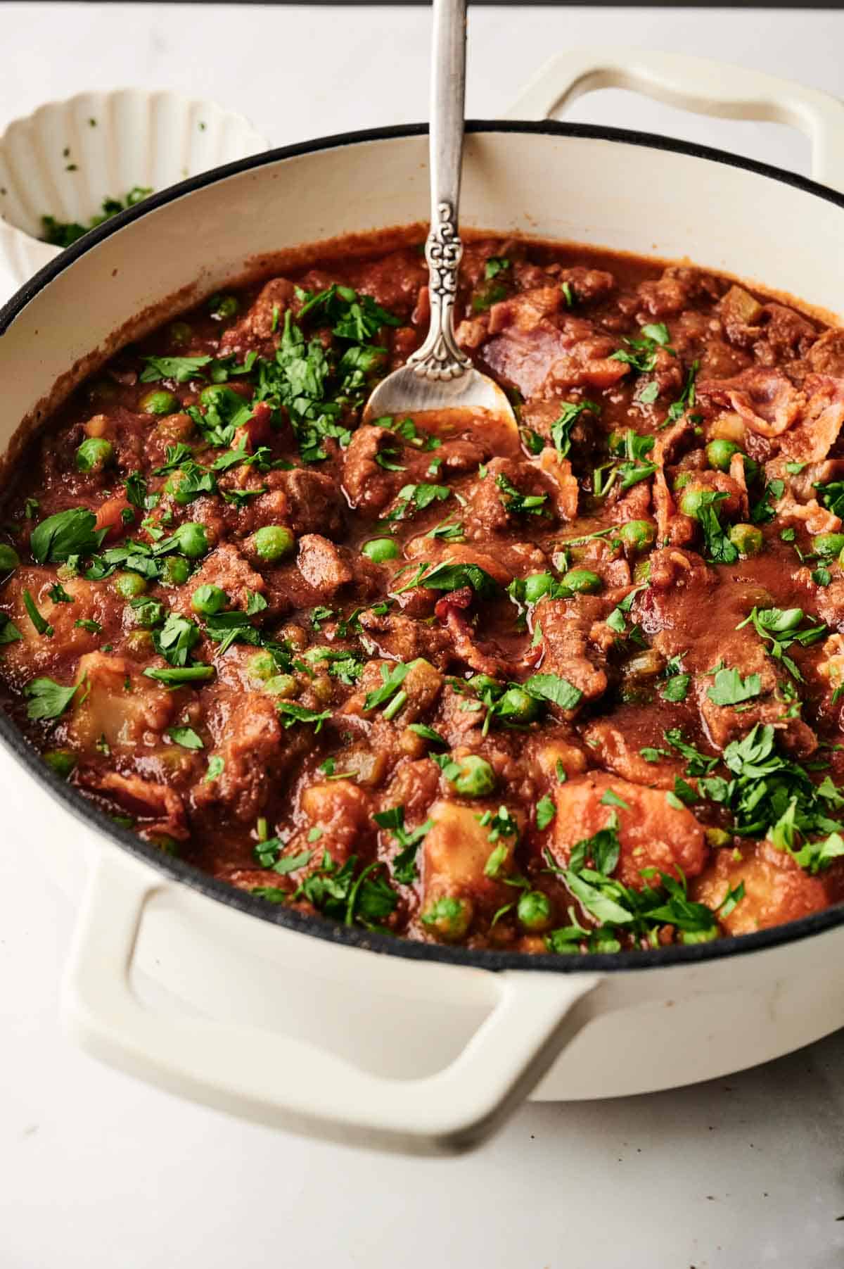 A white pot filled with a hearty beef stew, featuring chunks of meat, peas, carrots, and herbs, with a spoon resting inside.