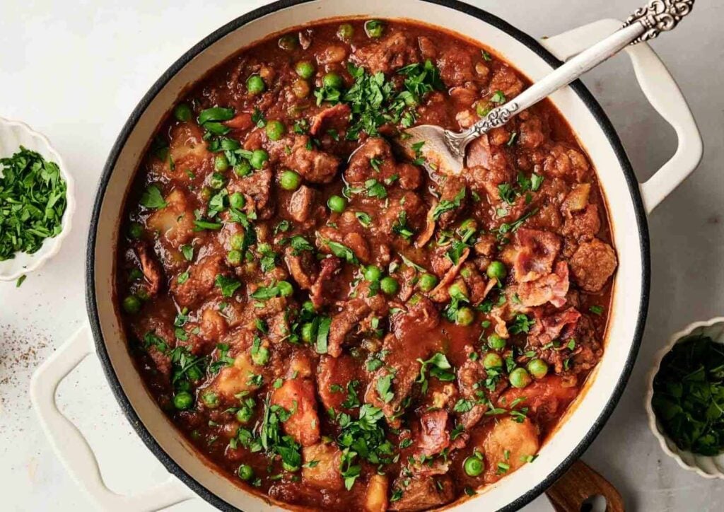 A pot of hearty beef stew brimming with peas, potatoes, carrots, and chopped herbs, with a spoon resting inside.